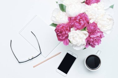 Kahve, cep telefonu, kağıt, kalem, bardak ve beyaz masa arkasında beyaz ve pembe şakayık çiçekleri olan iş yeri. Düz yatış, üst görünüm