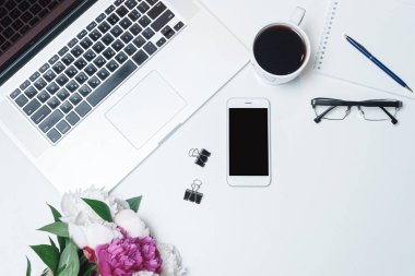 Dizüstü bilgisayarı, gözlükleri, defteri, cep telefonu ve beyaz masa arkasında pembe ve beyaz şakayık çiçekleri olan bir işyeri. Düz yatış, üst görünüm
