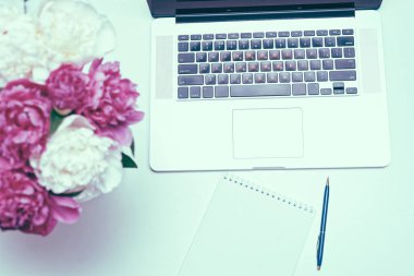 Beyaz masa arkasında defter, kalem, pembe ve beyaz şakayık çiçekli işyeri. Düz yatış, üst görünüm