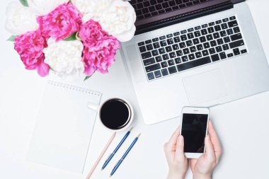 Dizüstü bilgisayarı, cep telefonu, bir fincan kahve ve kadın eli, defter, kalem ve beyaz fonda beyaz şakayık çiçekleri olan çalışma alanı. Düz yerde, üst tarafta. Serbest İşyeri