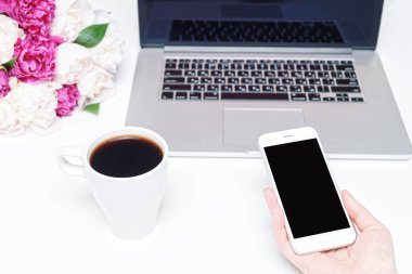 Dizüstü bilgisayarı, cep telefonu, bir fincan kahve ve beyaz arka planda kadın eli, not defteri ve pembe ve beyaz şakayık çiçekleri olan çalışma alanı. Düz yerde, üst tarafta. Serbest İşyeri