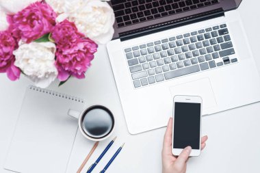 Dizüstü bilgisayarı, cep telefonu, bir fincan kahve ve kadın eli, defter, kalemler ve beyaz arka planda pembe ve beyaz şakayık çiçekleri olan çalışma alanı. Düz yerde, üst tarafta. Serbest İşyeri