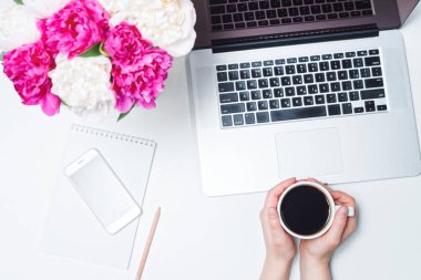 Çalışma alanı cep telefonu, dizüstü bilgisayar, bir fincan kahve ve kadın eli, defter, kalemler ve beyaz arka planda pembe ve beyaz şakayık çiçekleri. Düz yatış, üst görünüm, serbest çalışma yeri