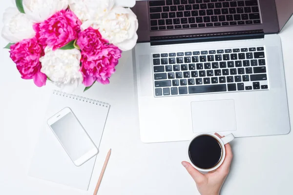 Workspace with laptop, mobile phone, cup of coffee and female hand, notebook, pens, and pink and white peony flowers on the white background. Flat lay, top view, Freelancer working place