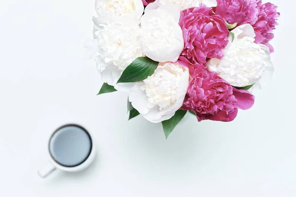 Working place with cup of coffee and white and pink peony flowers on white table background. Flat lay, top view