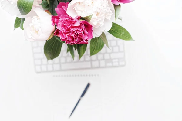 Workplace with keyboard, pen and pink and white peony flowers on the white table background. Flat lay, top view
