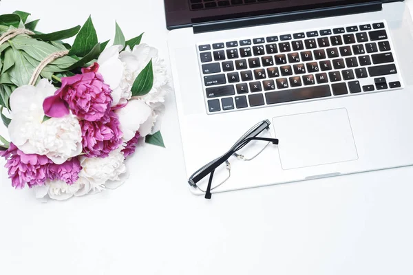 Workplace with laptop, glasses and pink and white peony flowers on the white table background. Flat lay, top view