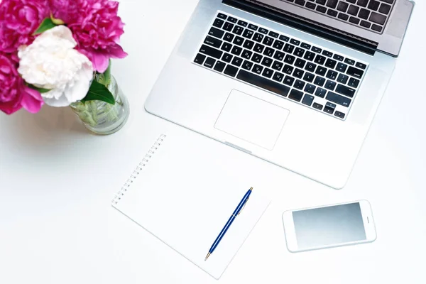 Workplace with laptop, notebook, pen and pink and white peony flowers on the white table background. Flat lay, top view