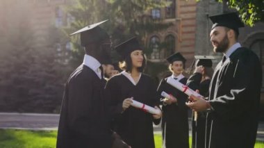 Genç ve güzel kadın ve yakışıklı erkeklerde önlük ve diploma tutarak ve sonra güneşli bir mezuniyet töreni Üniversitesi avlu üzerinde konuşmak kap. Karışık ırklar. Dışarıda
