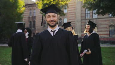 Portre yakışıklı genç mezunu siyah geleneksel elbise ve şapka kameraya poz ve Evet jest yapıyor. Arka planda mezunları. Açık