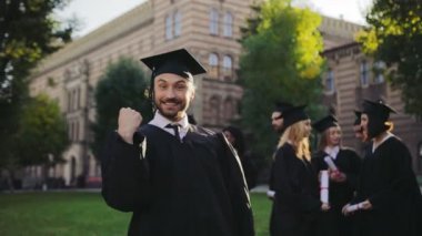 Mutlu neşeli ve komik adam geleneksel mezuniyet elbisesi ve kap eğleniyor ve kameraya ekşitmeden elinde diploma ile portre vurdu. Arka planda mezunları. Açık havada