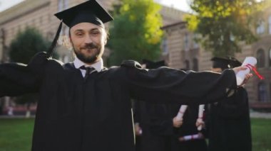 Kamera önünde duran ve parmağını pes neşeli çekici erkek mezunu. Arka planda mezunları. Açık havada. Portre vurdu