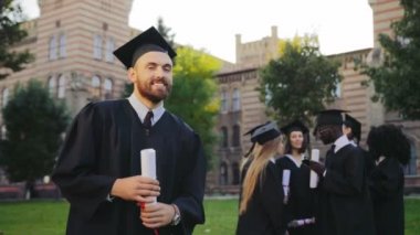 Portre çekici mezun gencin siyah geleneksel giysiler ve kameraya poz ve gülümseyen kap. Arka planda mezunları. Açık
