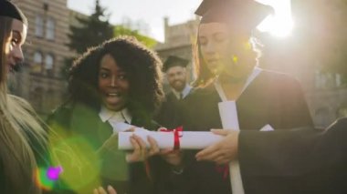 Çekici genç kadın mezunları geleneksel önlük ve diploma unwrapping ve Wow bakarak zaman söyleyerek kapaklar. Çocuklar arka planda. Dışarıda