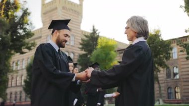 Orta yaşlı çekici mezun adam tebrik ediyor ve onu Üniversite binası önünde bir diploma sertifika veren Profesör. Diğer mezunlar arka planda. Açık