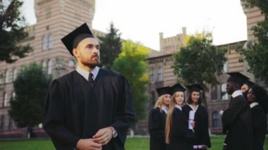 Portre çekim mezun gencin siyah geleneksel elbise ve diploma ile kamera önünde duran ve ellerini geçmek kap. Arka planda mezunları. Açık havada