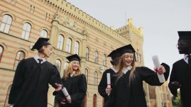Geleneksel siyah önlük ve bina ve neşeyle konuşurken Üniversitesi yürüyüş kapaklar ve Genç multi etnik mezunları grubu. Mezuniyet günü. Açık havada