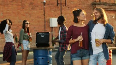 Çekici genç çift birbirine bakarak ve çatı partisi kameraya poz bira ile. Tuğla duvar ve dans eden insanlar arka plan. Kentsel alan. Açık. Multiethnical