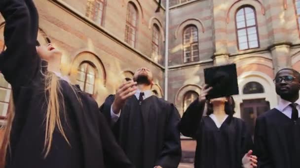 Felices graduados de carreras mixtas de pie en el patio de la Universidad y lanzando sus gorras de graduación en el aire uno por uno. Exterior — Vídeos de Stock