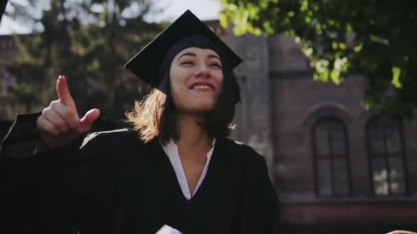 在毕业日, 有魅力的女毕业生在她的多族裔男性朋友中大声大笑。户外。特写 — 图库视频影像