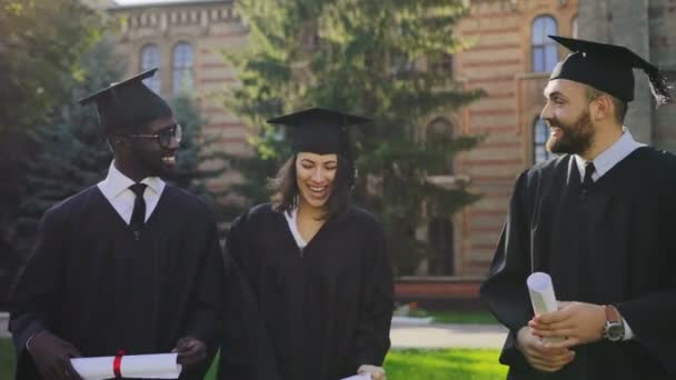 Amigos multi étnicos graduados caminando con sus diplomas y en ropa tradicional después de la ceremonia de graduación cerca de la Universidad. Al aire libre — Vídeo de stock