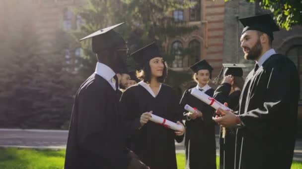 Mujer joven bonita y hombres guapos en batas y gorra con diplomas y hablando después de la ceremonia de graduación en el patio de la Universidad en un día soleado. Razas mixtas. Afuera. — Vídeo de stock