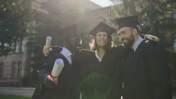 Retrato de las razas mixtas masculina y femenina en ropa tradicional y gorras posando a la cámara y abrazándose en su día de graduación. Exterior — Vídeos de Stock