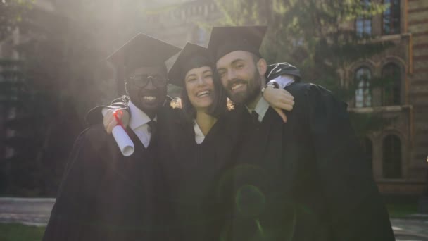 Retrato filmado da jovem mulher atraente e homens em roupas e bonés tradicionais posando para a câmera e abraçando em seu dia de formatura. Fora — Vídeo de Stock