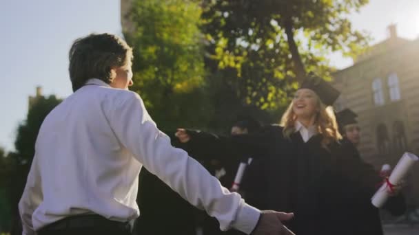 Blonde young woman in traditional gown and cap meeting her gray-haired man and hugging him after the graduation ceremony. Outdoor — Stock Video