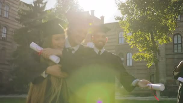 Os graduados alegres étnicos múltiplos que abraçam e congratulam um a outro alegremente com a graduação no dia ensolarado. Fora — Vídeo de Stock