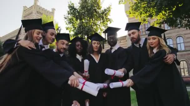 Carreras mixtas graduados en ropa tradicional de pie juntos y lanzando al aire sus diplomas. De cerca. Afuera. — Vídeo de stock