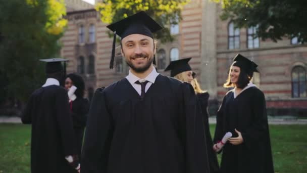 Retrato del apuesto joven graduado en vestido tradicional negro y gorra posando a la cámara y haciendo el gesto SI. Graduados en segundo plano. Exterior — Vídeo de stock