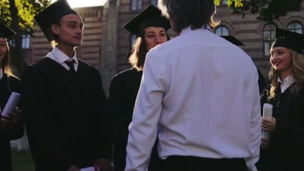 Parte trasera del hombre de pelo gris hablando alegremente con un hermoso graduado y felicitándola con el acabado del tachonado. Entre otros graduados. Afuera. — Vídeos de Stock