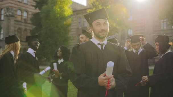 Porträt des gut aussehenden Mannes, der in traditioneller Promotionskleidung mit Diplom in der Hand steht und hoffnungsvoll in den Himmel blickt. Absolventen im Hintergrund. Außen — Stockvideo