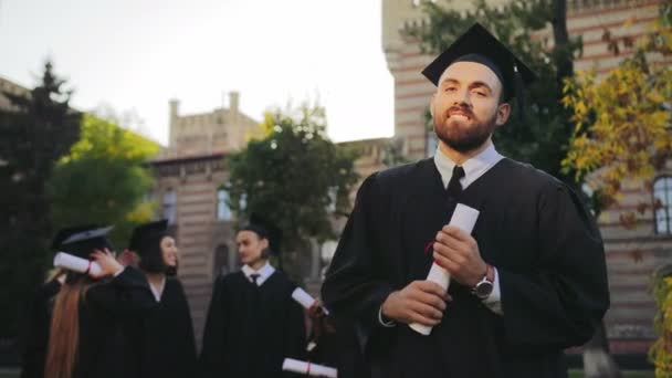 肖像拍摄的英俊的年轻毕业的黑色传统礼服和帽子摆在相机和微笑。毕业生的背景。外面 — 图库视频影像
