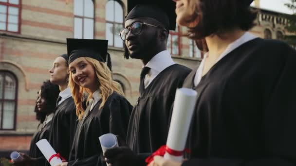 Multi etnische afgestudeerden in traditionele kleding staande in de lijn en houden van hun diploma's aan hun universiteit. Close-up. Uitzicht vanaf de kant. Buitenshuis — Stockvideo