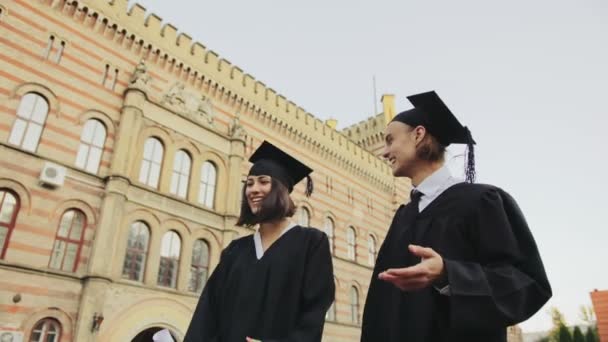 Ein paar junge Absolventen in traditionellen schwarzen Gewändern und Mützen spazieren in der Nähe des Universitätsgebäudes und unterhalten sich. Abi-Tag. Außenbereich — Stockvideo