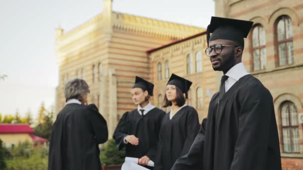 Portrait du diplômé afro-américain souriant posant devant la caméra et croisant les mains devant l'Université. Diplômés avec professeur en arrière-plan. Extérieur — Video