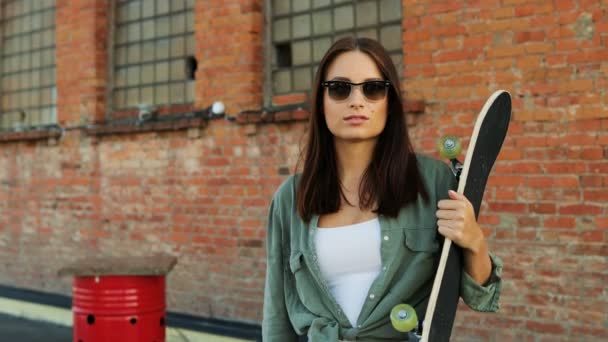 Portrait of beautiful young woman with a skateboard posing to the camera and laughing on the brick wall background. Outside — Stock Video