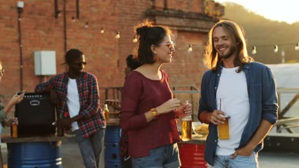 Atractiva pareja joven hablando, riendo y bebiendo cerveza en la fiesta de la azotea. La pared de ladrillo y el fondo del cielo. Espacio urbano. Al aire libre. Multiétnico — Vídeos de Stock