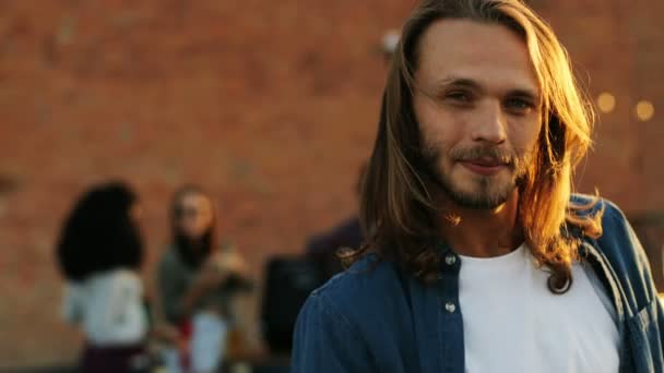 Tiro de perto de homem bonito com cabelo loiro longo sorrindo direto para a câmera na festa no telhado. O fundo da parede de tijolos. Espaço urbano. Lá fora. Retrato — Vídeo de Stock