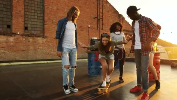 Giovani uomini che insegnano giovane ragazza come fare skateboard alla festa sul tetto. Mi diverto. Il muro di mattoni e divertente sfondo ragazze. Fuori. Razze miste — Video Stock