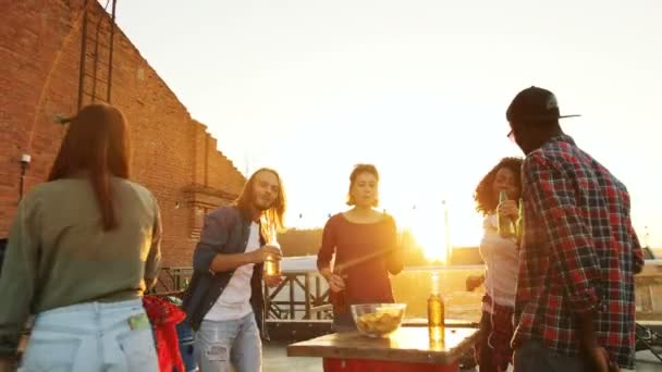 Baráti fiatal táncoló és csinálás pirítós sört ételek a tetőtéri párt az asztal körül naplemente. A téglafal háttér. Kültéri. Többnemzetiségű — Stock videók