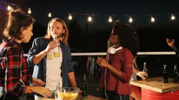 Jovens multiétnicos conversando e bebendo cerveja ao redor das mesas na festa do telhado à noite. O fundo da parede de tijolos. Fora . — Vídeo de Stock