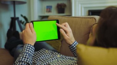 Rengarenk gömlek ve kanepede yatan ve siyah tablet bilgisayar onun elinde yeşil ekranda kaydırma kot erkekte omzuna üzerinde görüntüleyin. Chroma anahtar. Evde istirahat. Kapalı