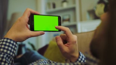 Rengarenk gömlek ve kanepede yatan ve onun elinde siyah smartphone yeşil ekranda kaydırma kot erkekte omzuna üzerinde görüntüleyin. Chroma anahtar. Evde istirahat. Yakın çekim. İçinde