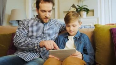 Orta portre çekim baba ve küçük oğul birlikte kanepede, holding bir tablet bilgisayar ve onlar oynamak her iki çalışan çocuk oturan yaşlı. Kapalı