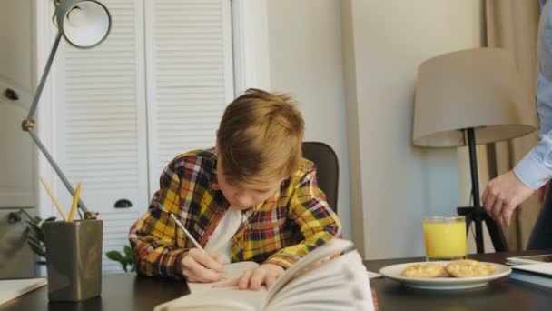 Bello il padre che viene al tavolo dove suo figlio scrive i compiti nella bella stanza accogliente di casa. Succo e biscotti sul tavolo. Dentro. — Video Stock