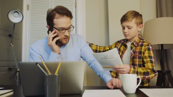 Padre ocupado trabajando en el ordenador portátil en la mesa de la oficina y hablando por teléfono cuando su hijo pequeño viene a él y pidiendo ayuda con la tarea. Papá se niega a su hijo. En casa. Adentro. — Vídeo de stock