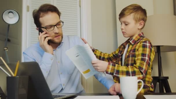 Papá ocupado en gafas sentado en la computadora portátil en el escritorio y hablando por teléfono cuando su hijo pequeño viene y pide ayuda con la tarea. Padre lo envió fuera. En casa. De interior — Vídeos de Stock